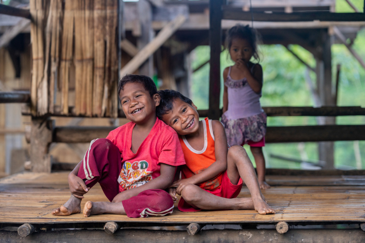 one boy leans on another boy smiling on a wooden mat in an outdoor setting