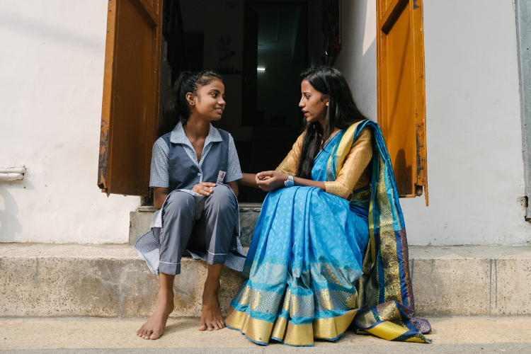 a younger Indian girl sits on a stoop in front of an open door talking to an older Indian woman